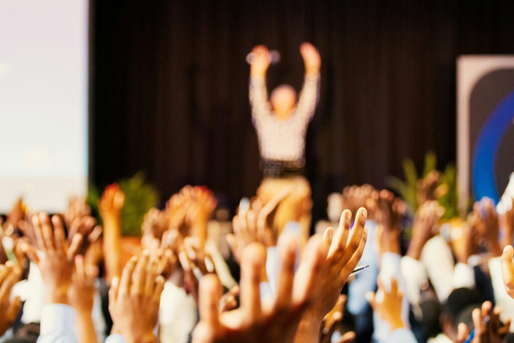 Un évènement avec une salle de personnes levant de mains en air devant un personne sur scene avec ses mains levées en air